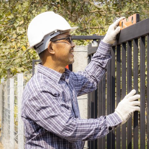 Installer in a white hard hat and work gloves holding a level on top of a black metal picket fence