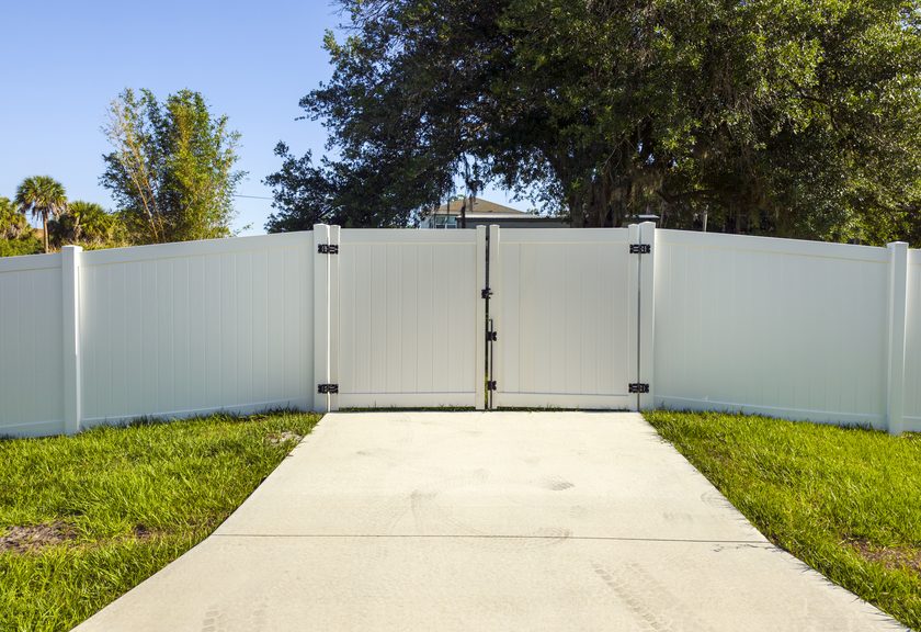White vinyl privacy fence with a double gate across a concrete walk, palms and trees behind